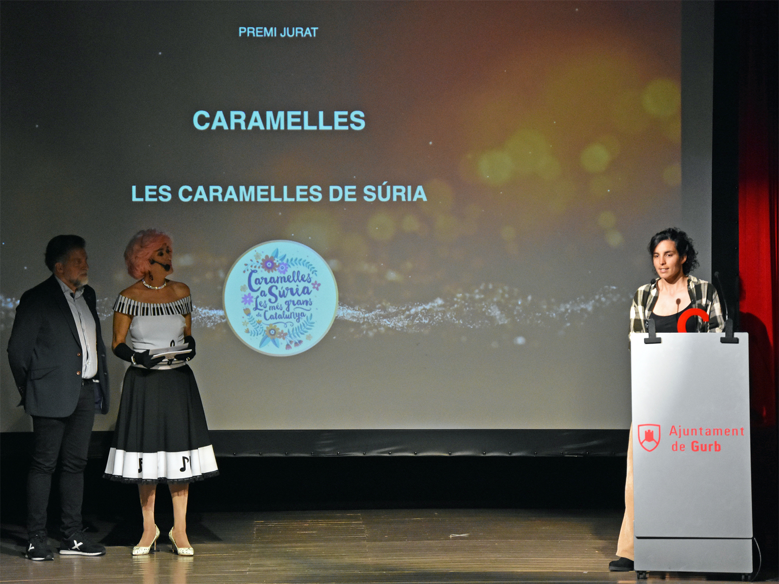 La regidora de Festes, Alba Martínez, parla després de recollir el Premi Josep Anselm Clavé del Cant Coral a Catalunya, al costat del president de la Federació d'Ateneus de Catalunya, Pep Morella, i de la presentadora de l'acte, Brigitta Lamoure.